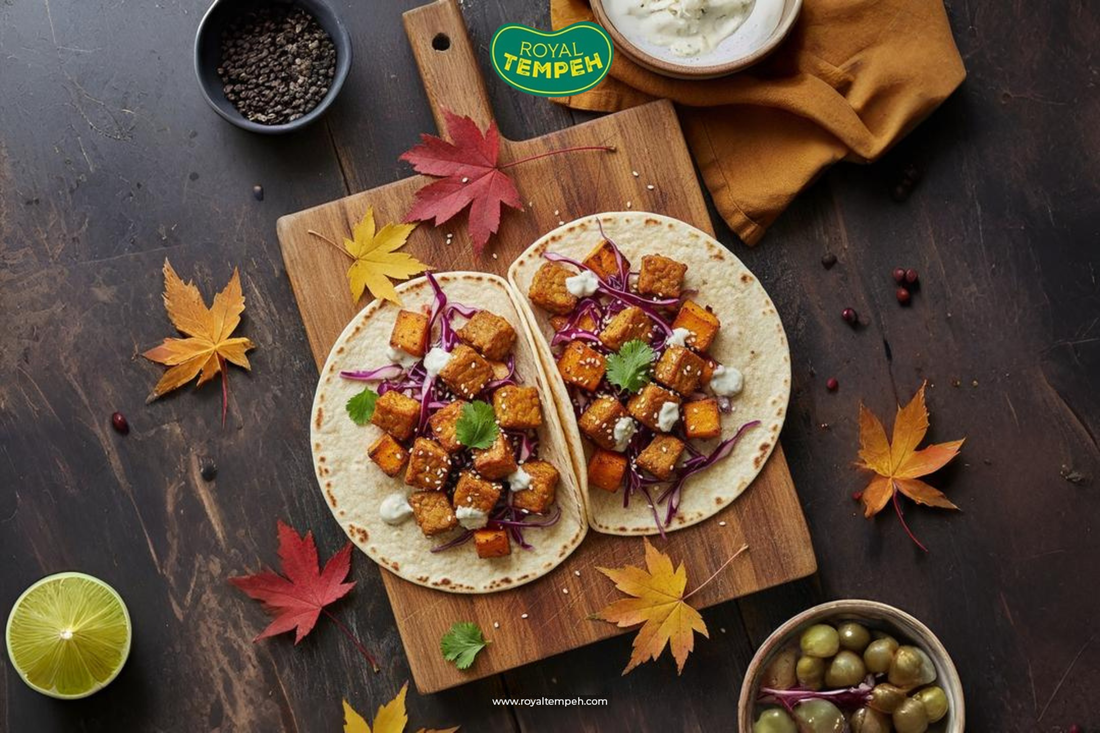 Two soft tacos filled with roasted tempeh cubes, red cabbage, and sauce on a wooden board surrounded by autumn leaves and lime, with the Royal Tempeh logo above.