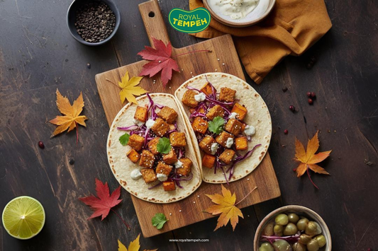 Two soft tacos filled with roasted tempeh cubes, red cabbage, and sauce on a wooden board surrounded by autumn leaves and lime, with the Royal Tempeh logo above.
