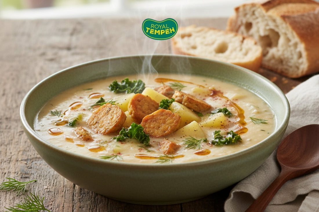 Bowl of creamy soup with tempeh slices, potatoes, and greens, garnished with herbs and served with crusty bread on a rustic wooden table, featuring the Royal Tempeh logo above.