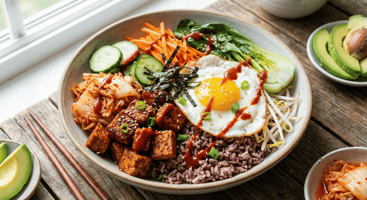 A colorful rice bowl topped with tempeh, kimchi, cucumbers, carrots, bok choy, bean sprouts, seaweed strips, and a sunny-side-up egg drizzled with sauce.