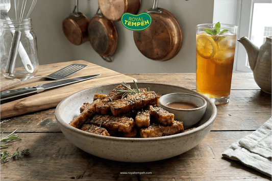 Plate of glazed tempeh slices topped with sesame and herbs served with dipping sauce on a rustic wooden table.