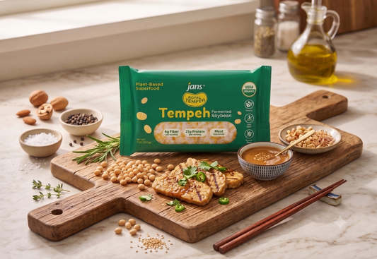 A professional wholesale product display showcasing a green package of Jans Royal Tempeh on a rustic wooden cutting board alongside grilled tempeh slices, a dish of dipping sauce, a small bowl of soybeans, and chopsticks.