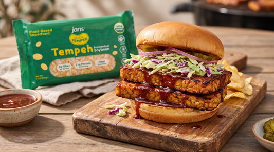 A stacked BBQ tempeh sandwich layered with barbecue sauce, crunchy coleslaw, and pickles on a wooden cutting board next to potato chips, barbecue sauce, and a green package of Jans Royal Tempeh Block.