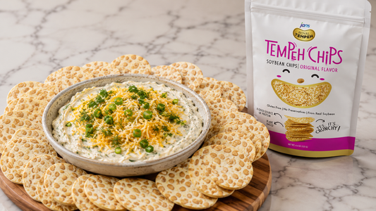 A ceramic bowl of creamy boat dip garnished with shredded cheddar cheese and sliced green onions, surrounded by round Jans Tempeh Chips on a wooden platter next to a bag of Jans Tempeh Chips.