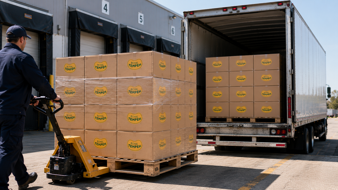 A warehouse worker using a yellow hand pallet jack to move a pallet of cardboard shipping boxes bearing yellow Royal Tempeh logos into the back of a commercial semi-truck.