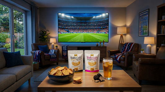 A modern living room set up for a soccer watch party, featuring a large TV streaming a match over a coffee table displaying bags of Jans Tempeh Chips, a bowl of crisps, and a cold drink.