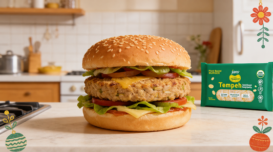 A thick, grilled plant-based burger patty stacked inside a sesame seed bun with melted cheese, lettuce, and tomato, sitting on a kitchen counter next to a green package of Royal Tempeh Burger.