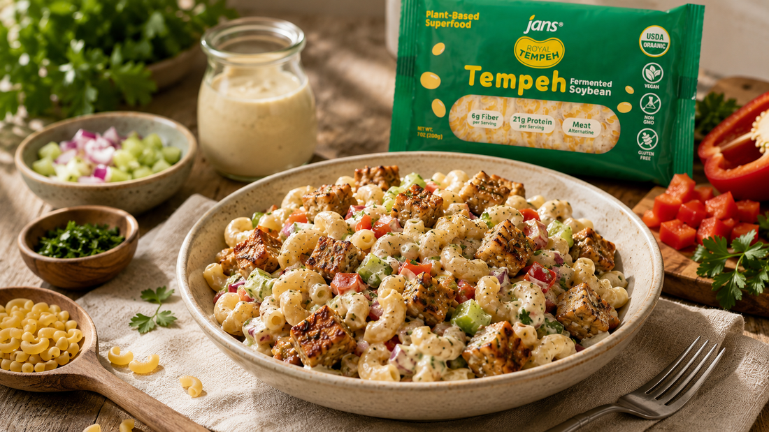 A large white ceramic bowl filled with a creamy macaroni salad mixed with chopped vegetables and pan-seared tempeh cubes, sitting on a wooden counter next to a green package of Royal Tempeh, a jar of dressing, and a wooden spoon.