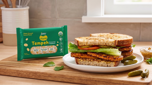 A thick, grilled deli sandwich layered with thinly sliced marinated tempeh, lettuce, and tomato slices on toasted artisan bread, displayed on a wooden board next to a green package of Jans Royal Tempeh and small pickles.