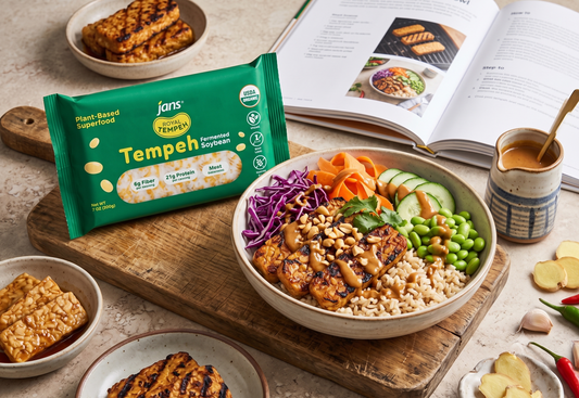 A wholesome grain bowl filled with grilled tempeh cubes, shredded purple cabbage, carrots, edamame, and cucumbers drizzled with peanut sauce, displayed next to a green package of Jans Royal Tempeh and an open cookbook.