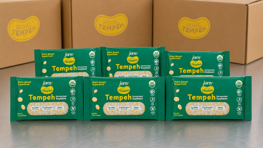 Seven green packages of Royal Tempeh neatly displayed on a metallic countertop in front of three brown cardboard wholesale shipping boxes featuring yellow Royal Tempeh circular logos.