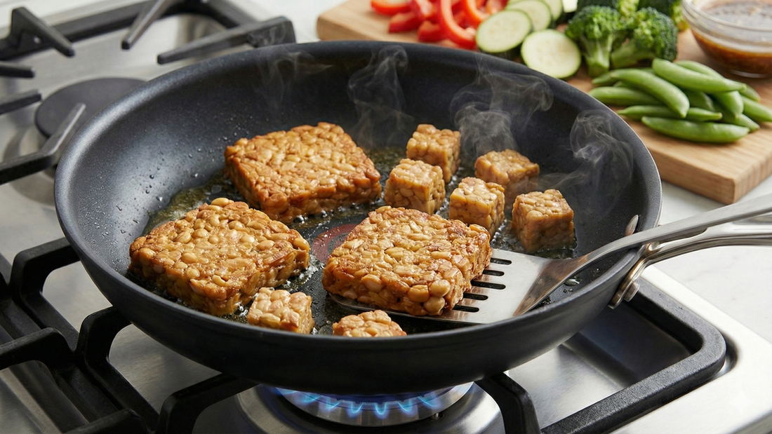 Thick, square slabs and smaller cubes of Royal Tempeh being pan-fried to a golden-brown finish in a skillet on a gas stove, with fresh broccoli, peppers, and zucchini on a cutting board in the background.