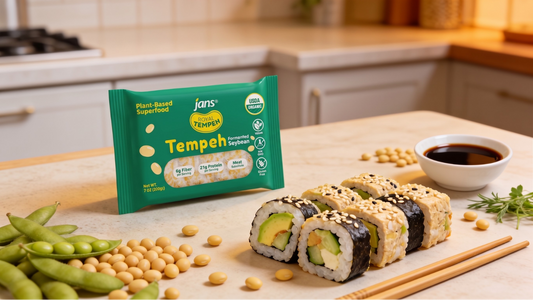 Four slices of sesame-crusted tempeh sushi rolls filled with avocado and cucumber, displayed on a wooden table next to a green package of Royal Tempeh, a small bowl of soy sauce, loose soybeans, and fresh edamame pods.