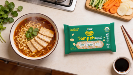 A steaming bowl of ramen noodles in a deep brown savory broth topped with four rectangular slices of pan-seared tempeh, placed next to a green package of Royal Tempeh and a cutting board with fresh vegetables.