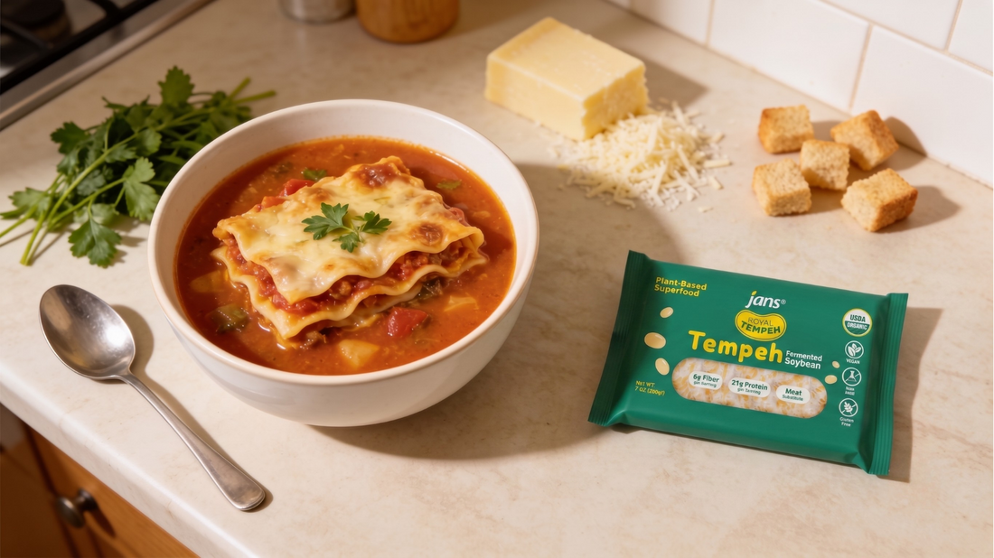 A white bowl filled with thick lasagna soup topped with melted mozzarella and fresh parsley, displayed on a kitchen counter beside a green package of Royal Tempeh, a spoon, garlic croutons, and a block of cheese.