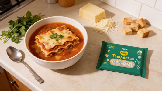 A white bowl filled with thick lasagna soup topped with melted mozzarella and fresh parsley, displayed on a kitchen counter beside a green package of Royal Tempeh, a spoon, garlic croutons, and a block of cheese.