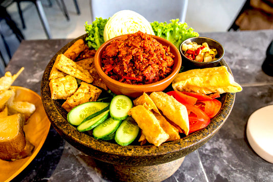 Traditional Indonesian platter with spicy sambal in the center, surrounded by tempeh, tofu, cucumber slices, tomatoes, cabbage, and lettuce.