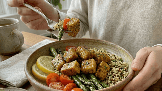 A close-up of a hand lifting a piece of lemon pepper tempeh with a fork from a bowl filled with quinoa, asparagus, cherry tomatoes, and lemon slices.