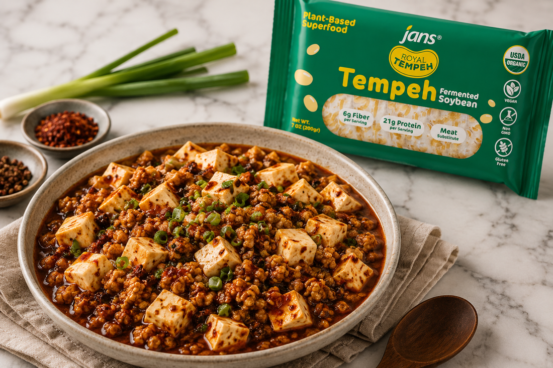 A oval ceramic bowl filled with fiery red Sichuan Mapo Tofu topped with minced green onions and crumbled tempeh, set on a kitchen counter next to a green package of Royal Tempeh, a small dish of red chili flakes, and scallions.