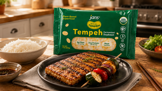 	A green package of Jans Royal Tempeh stands upright on a wooden kitchen counter behind a black plate featuring a beautifully grilled, sliced tempeh block and a colorful vegetable skewer, next to a bowl of white rice.