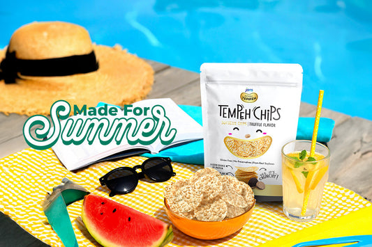 A white stand-up pouch of Jans Tempeh Chips Truffle flavor displayed next to a pool on a yellow checked towel, alongside a wooden bowl of chips, fresh watermelon slices, sunglasses, and a cold summer drink with a straw under a "Made For Summer" graphic.