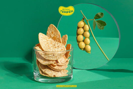 A clear glass filled with golden, crispy Royal Tempeh Chips displayed against a vibrant green background next to a round mirror reflecting a fresh green soybean pod under a yellow Royal Tempeh logo.