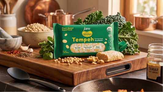A green retail pack of Jans Royal Tempeh Fermented Soybean displayed upright on a thick wooden cutting board in a warm kitchen kitchen, surrounded by fresh kale, a bowl of grains, garlic cloves, and kitchen pots.