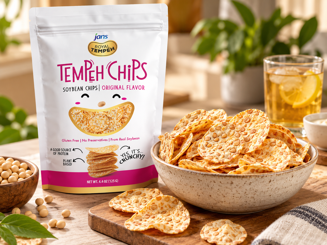 A bag of Jans Royal Tempeh Chips in Original Flavor next to a bowl of crunchy soybean chips, small bowls of soybeans, and glasses of iced tea on a warm wooden table.