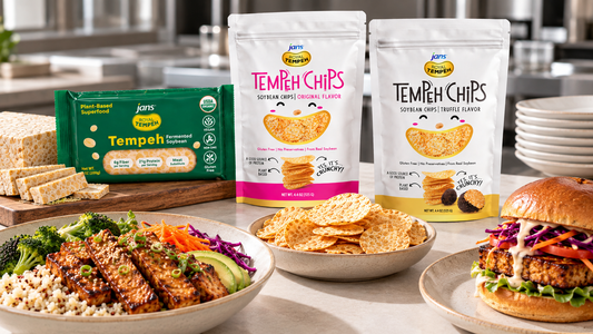 A product display featuring packaged Royal Tempeh, Original and Truffle flavor Tempeh Chips, and prepared plant-based dishes including a grain bowl, chip bowls, and a gourmet burger on a table in a modern commercial kitchen.