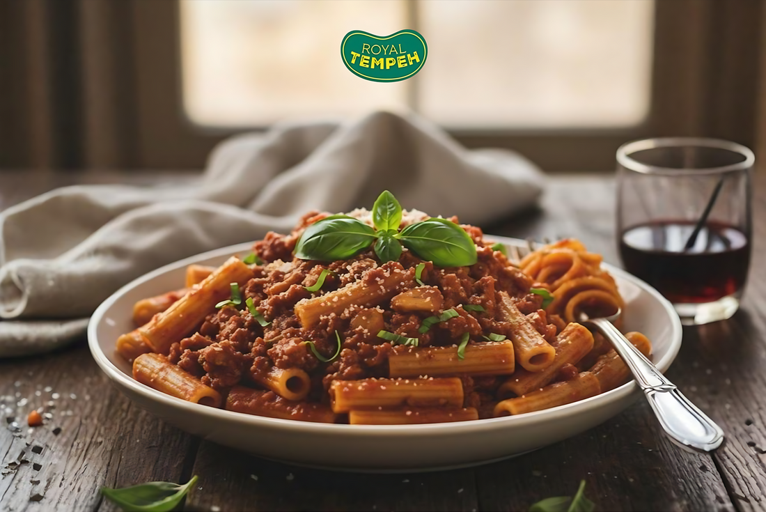 Plate of rigatoni pasta topped with tempeh bolognese sauce and fresh basil leaves, served on a rustic wooden table with the Royal Tempeh logo above.