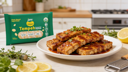 A white plate stacked with thin, glazed, golden-brown strips of marinated tempeh, presented next to a green package of Jans Royal Tempeh, fresh herbs, and lemon slices in a kitchen setting.