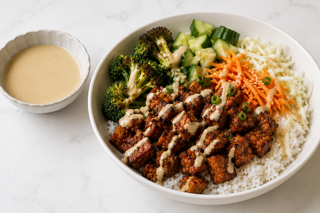 A white ceramic bowl filled with fluffy rice, steamed broccoli, diced cucumbers, shredded carrots, and golden, cubed Royal Tempeh drizzled with a rich sauce, served next to a small side dish of creamy tahini dressing.