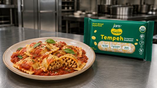 A plate of stuffed manicotti pasta topped with marinara sauce, fresh basil, and grated parmesan cheese sits on a stainless steel kitchen table next to a green package of Royal Tempeh.