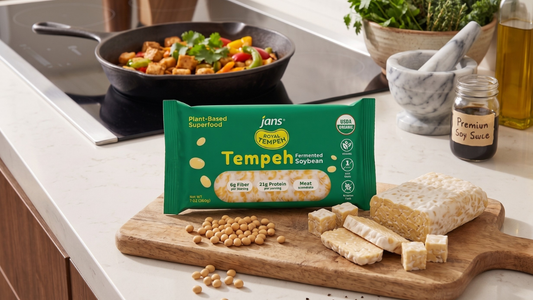 A retail pack of green Jans Royal Tempeh Organic Fermented Soybean on a wooden cutting board beside a raw, sliced tempeh block, loose soybeans, and a hot stir-fry cooking in a skillet.