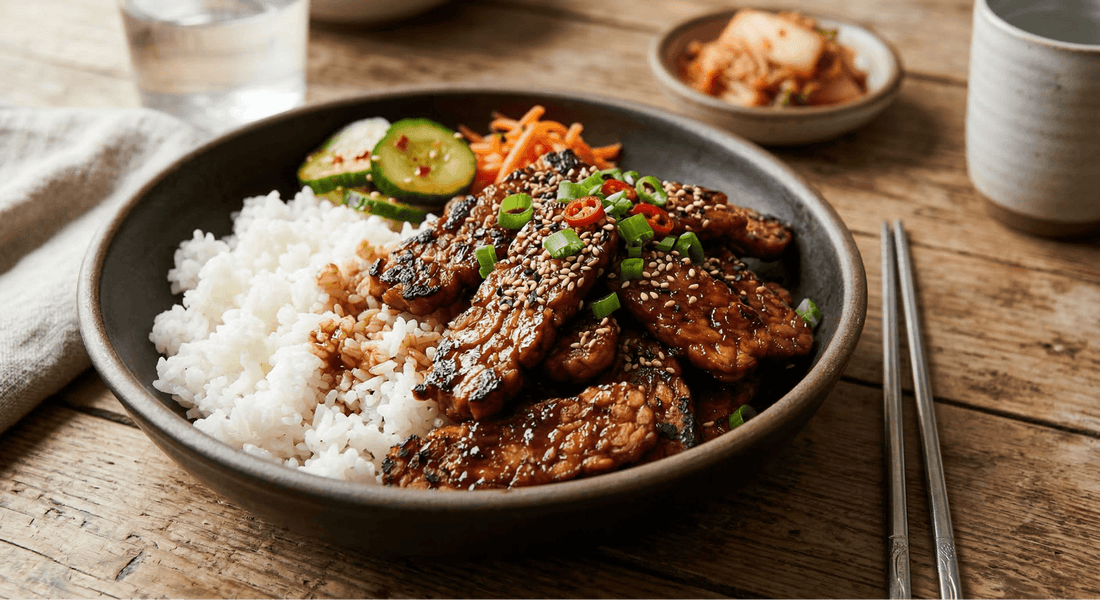 A bowl of savory Korean BBQ tempeh recipe featuring thinly sliced Royal Tempeh glazed in a dark bulgogi sauce, served with white rice, sesame seeds, and fresh cucumbers.