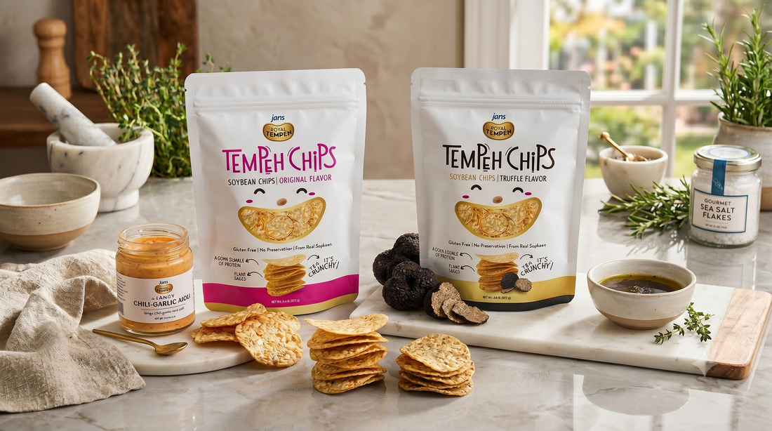 Two white stand-up pouches of Royal Tempeh Chips a pink Spicy Chili Garlic pack and a grey Truffle flavor pack, displayed on a marble counter next to scattered round, crispy chips, whole black truffles, and a jar of chili paste.