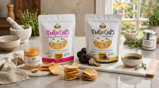 Two white stand-up pouches of Royal Tempeh Chips a pink Spicy Chili Garlic pack and a grey Truffle flavor pack, displayed on a marble counter next to scattered round, crispy chips, whole black truffles, and a jar of chili paste.