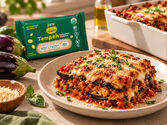 A plate of freshly baked tempeh eggplant parmesan garnished with fresh herbs, accompanied by a green package of Royal Tempeh and a baking dish in the background.