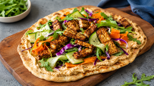 A toasted flatbread on a wooden cutting board generously spread with hummus and loaded with pan-seared Royal Tempeh strips, shaved carrots, cucumber, and fresh greens.