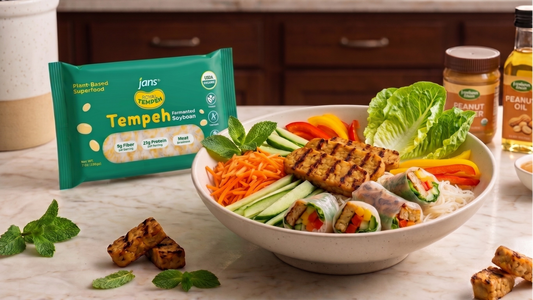 A wide white bowl filled with vermicelli rice noodles, pan-seared golden tempeh strips, shredded carrots, sliced cucumbers, and fresh mint, sitting next to a green package of Jans Royal Tempeh and bottles of peanut oil.