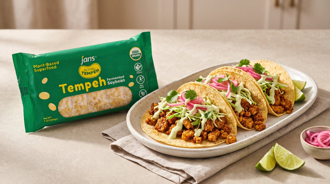 Three soft corn tacos filled with seasoned crumbled tempeh, shredded cabbage, zesty lime crema, cilantro, and bright pink pickled onions on a plate beside a package of Jans Royal Tempeh Fermented Soybean.