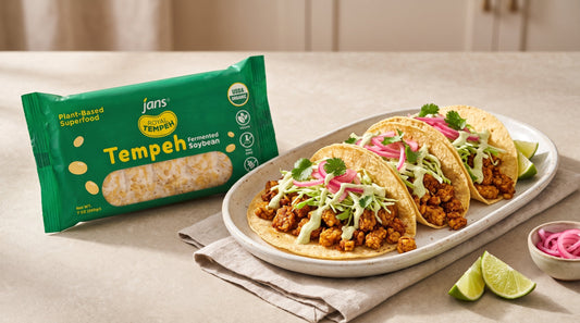 Three soft corn tacos filled with seasoned crumbled tempeh, shredded cabbage, zesty lime crema, cilantro, and bright pink pickled onions on a plate beside a package of Jans Royal Tempeh Fermented Soybean.