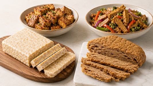 A wooden cutting board with a sliced raw tempeh block and a white marble board with sliced seitan, arranged in front of a ceramic bowl of cooked savory tempeh cubes and a fresh vegetable seitan stir-fry.