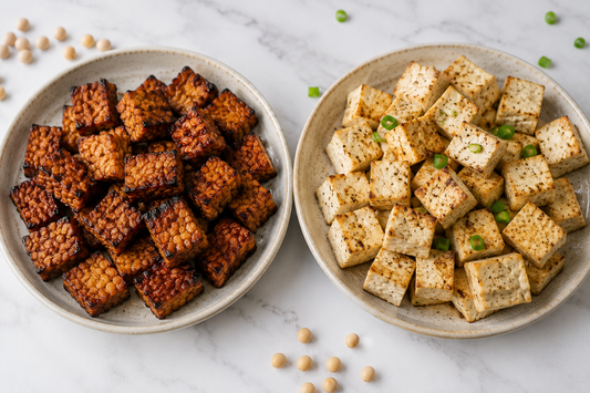 Two ceramic plates side by side on a light marble counter; the left plate holds deeply browned, seasoned cubes of cooked Royal Tempeh, while the right plate holds lighter, pan-seared cubes of seasoned tofu garnished with herbs.