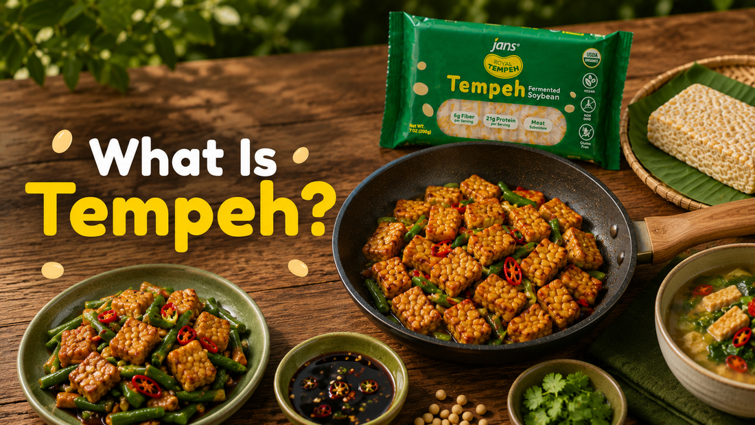 A black skillet filled with golden-brown, cubed cooked tempeh sits on a wooden table next to a green package of Jans Tempeh and small side dishes, with a bold graphic overlay that reads "What Is Tempeh?".