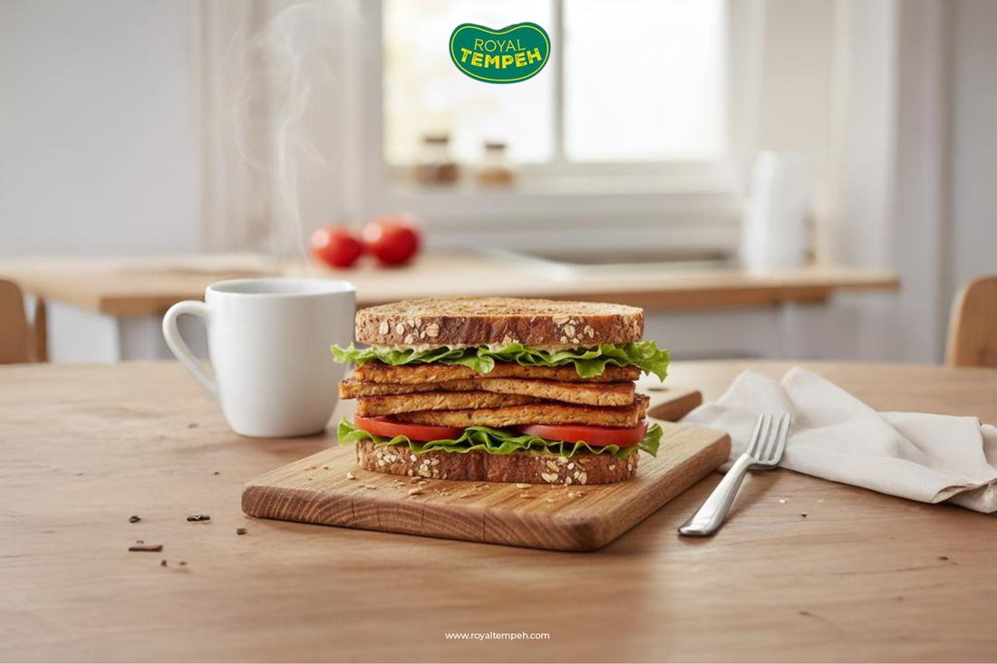 Royal Tempeh sandwich stacked with lettuce and tomato on whole grain bread, served on a wooden board with a hot cup of coffee.