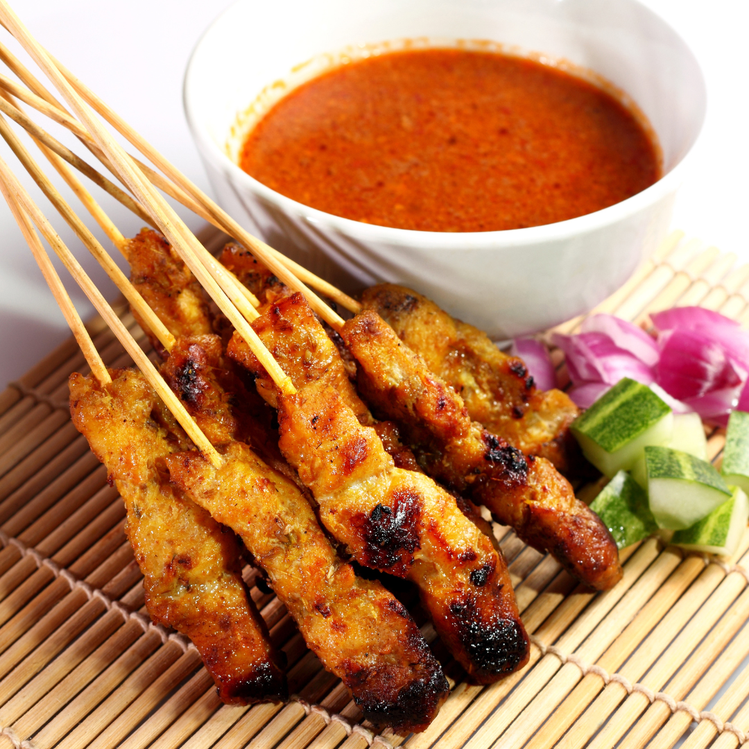 A bundle of grilled, golden-brown tempeh satay skewers arranged neatly on a bamboo mat next to a white bowl filled with rich, creamy peanut sauce, fresh cucumber slices, and red onions.