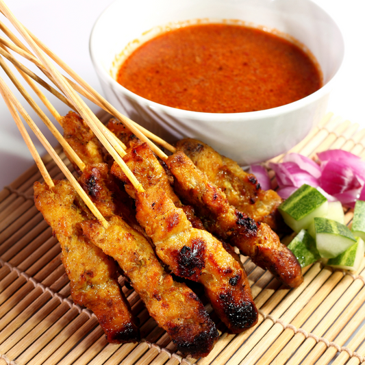 A bundle of grilled, golden-brown tempeh satay skewers arranged neatly on a bamboo mat next to a white bowl filled with rich, creamy peanut sauce, fresh cucumber slices, and red onions.