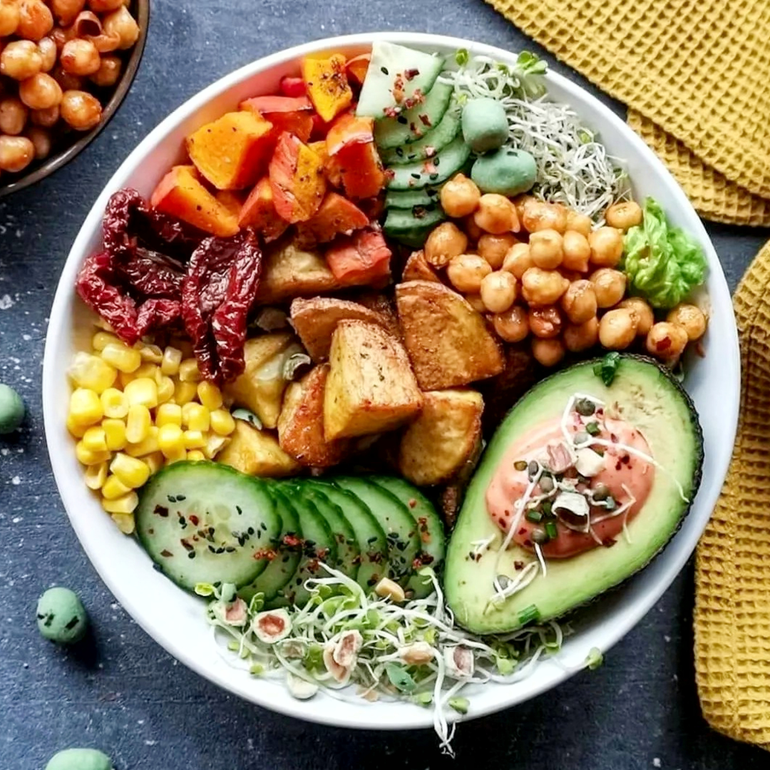A colorful plant-based buddha bowl viewed from above, packed with sliced cucumbers, avocado halves topped with microgreens, sweet corn, roasted potatoes, chickpeas, and mixed greens on a dark tabletop next to a yellow napkin.