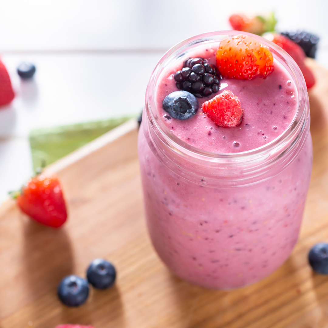 A thick, vibrant purple Tempeh Very Berry Smoothie served in a clear glass mason jar, topped with fresh strawberries, blueberries, and blackberries, resting on a wooden board.
